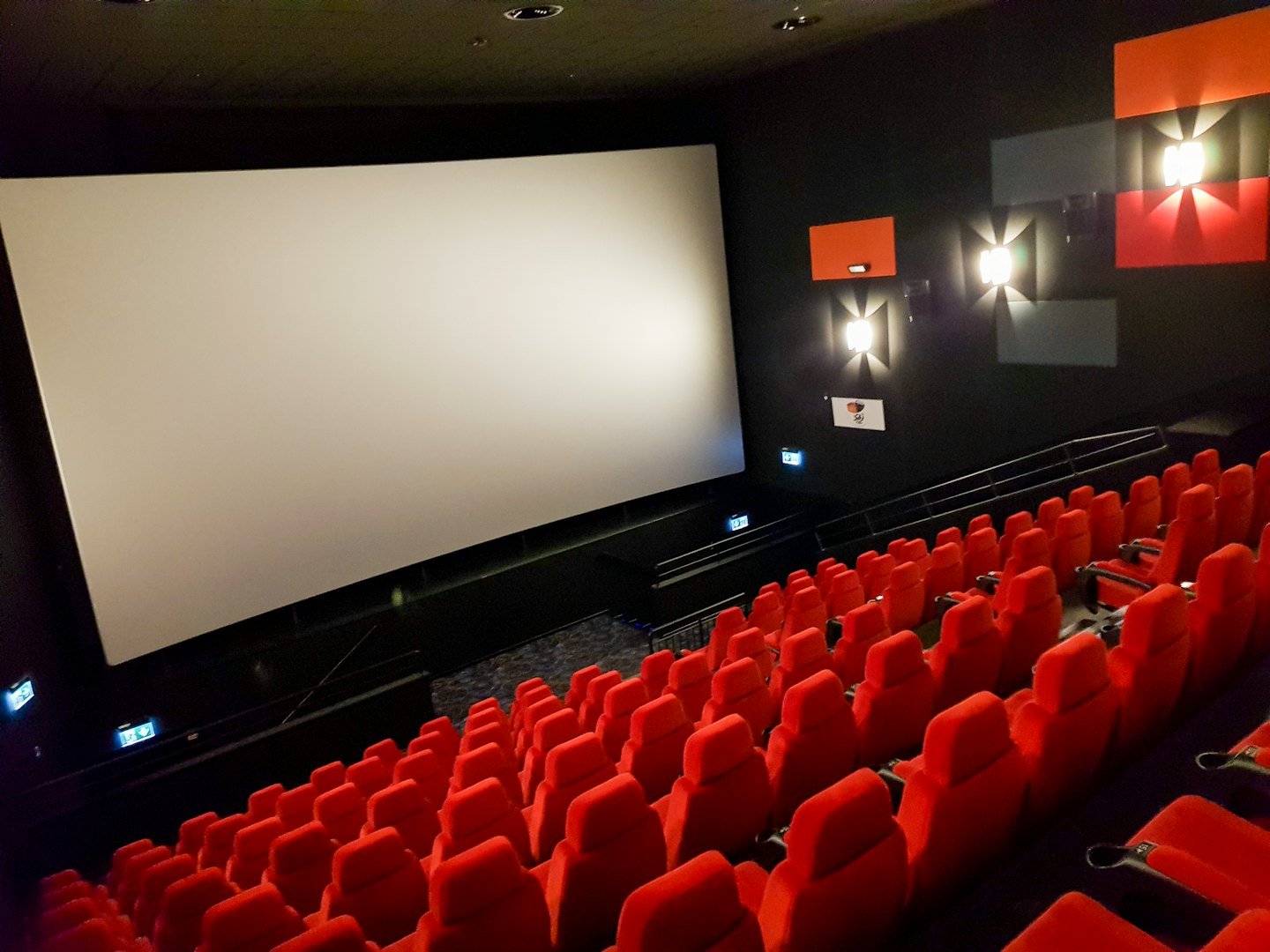 Empty cinema hall with rows of red seats and a white screen.